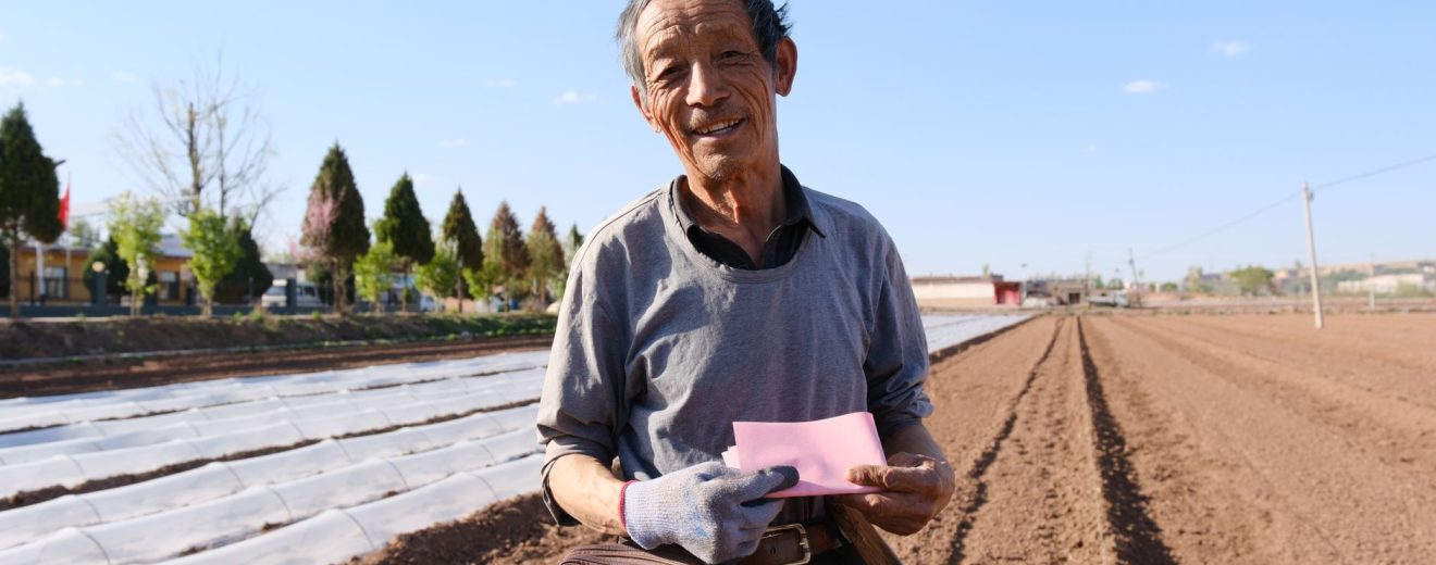 Agricultor idoso segura um documento e uma pá em uma área rural ensolarada.