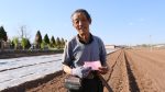 Agricultor idoso segura um documento e uma pá em uma área rural ensolarada.