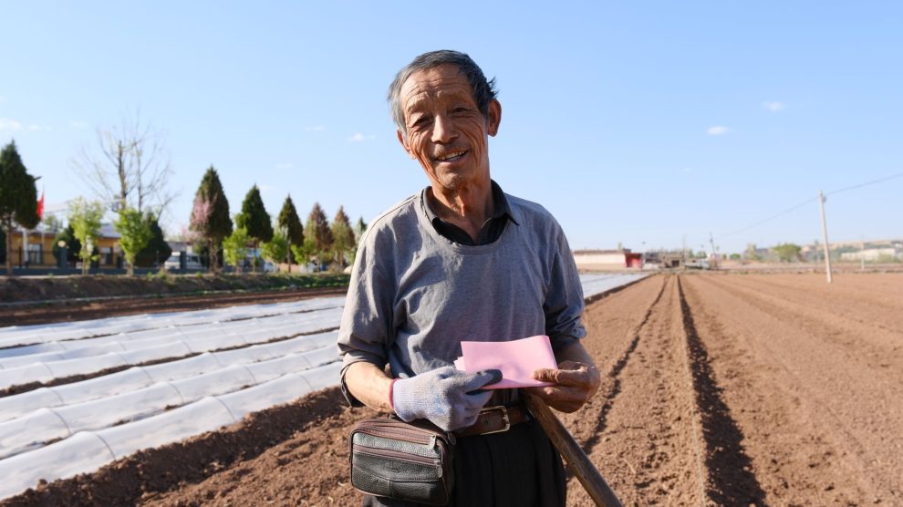Agricultor idoso segura um documento e uma pá em uma área rural ensolarada.